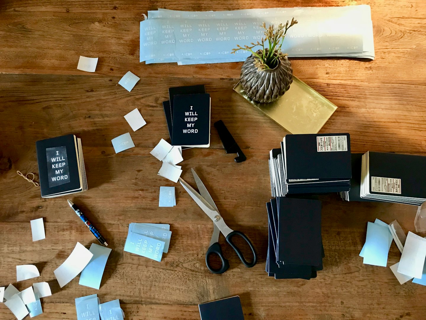 Notebooks being assembled with 'I WILL KEEP MY WORD' stickers on a wooden workbench.